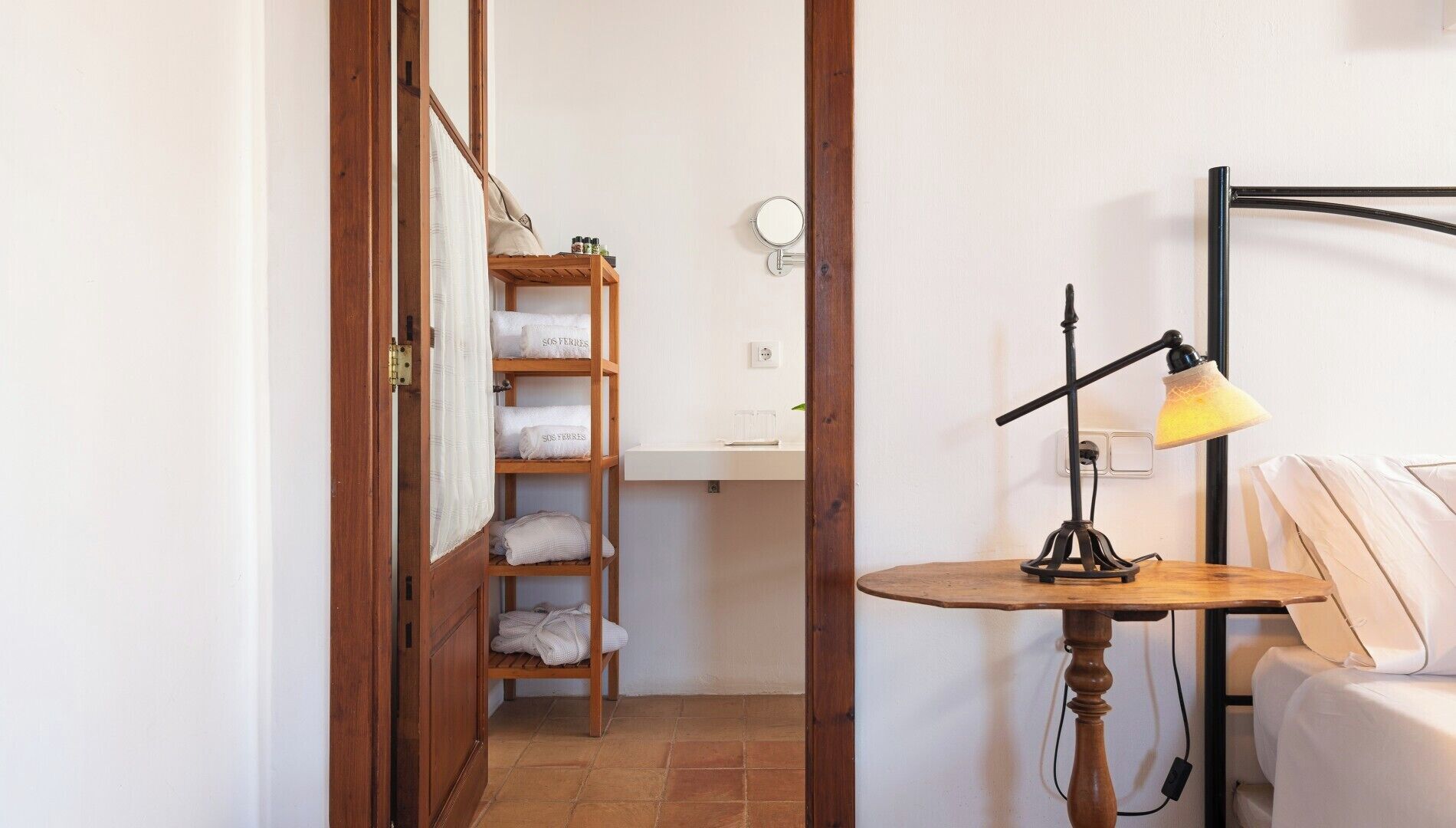 A small bathroom with a wooden shelf holding towels, a mirror, and a sink. There is a table with a lamp and a bed with white bedding.