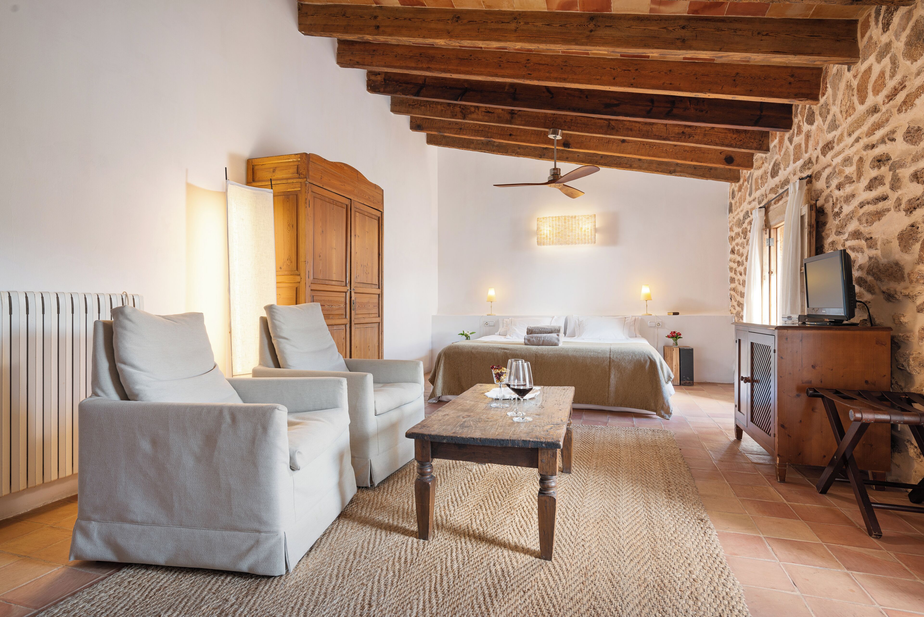 A cozy room with a stone wall, a wooden ceiling, a bed, a sofa, and a TV on a wooden cabinet.