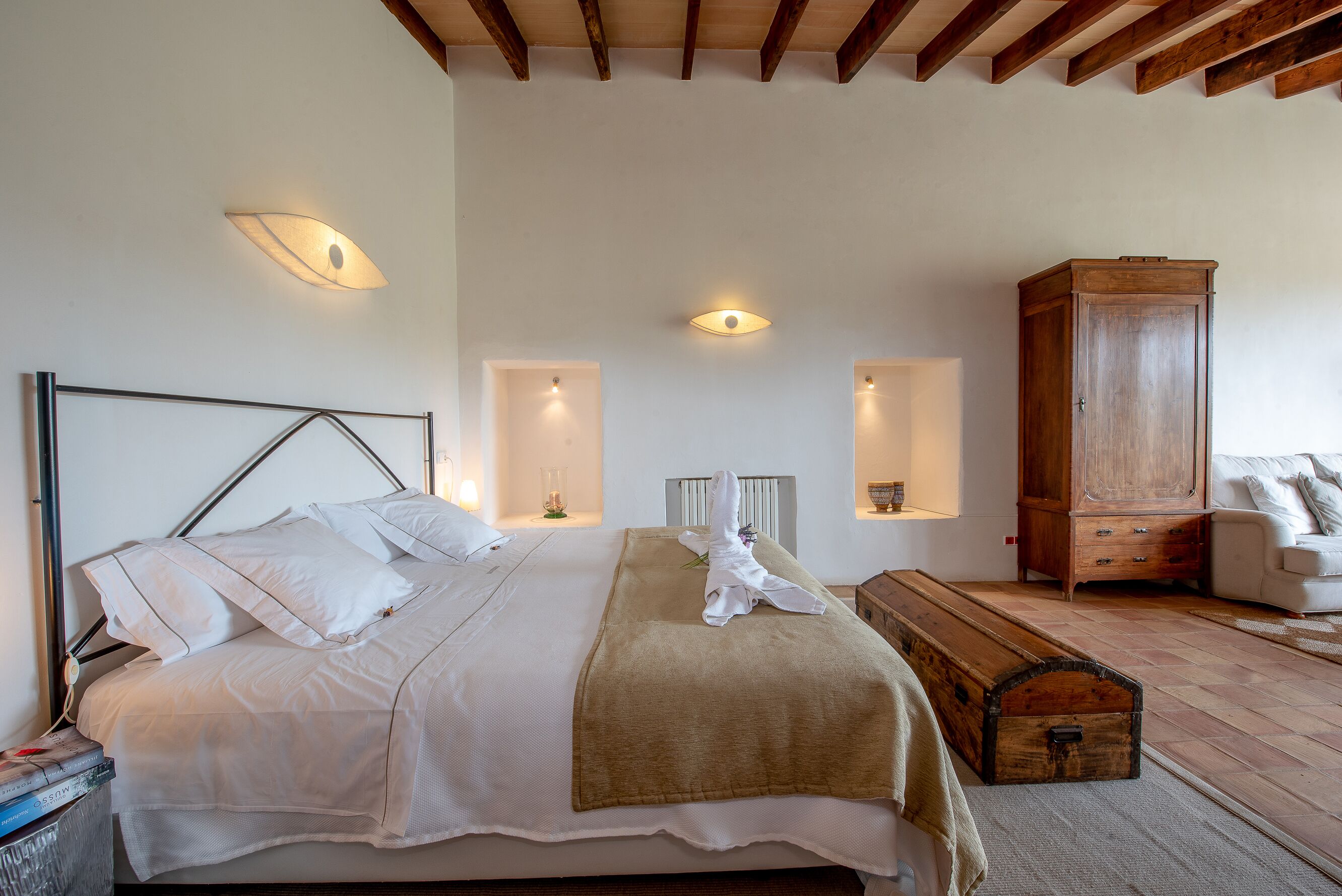 A neatly made bed with white linens, a wooden bench, and a vintage trunk in a room with exposed wooden beams and built-in wall sconces.