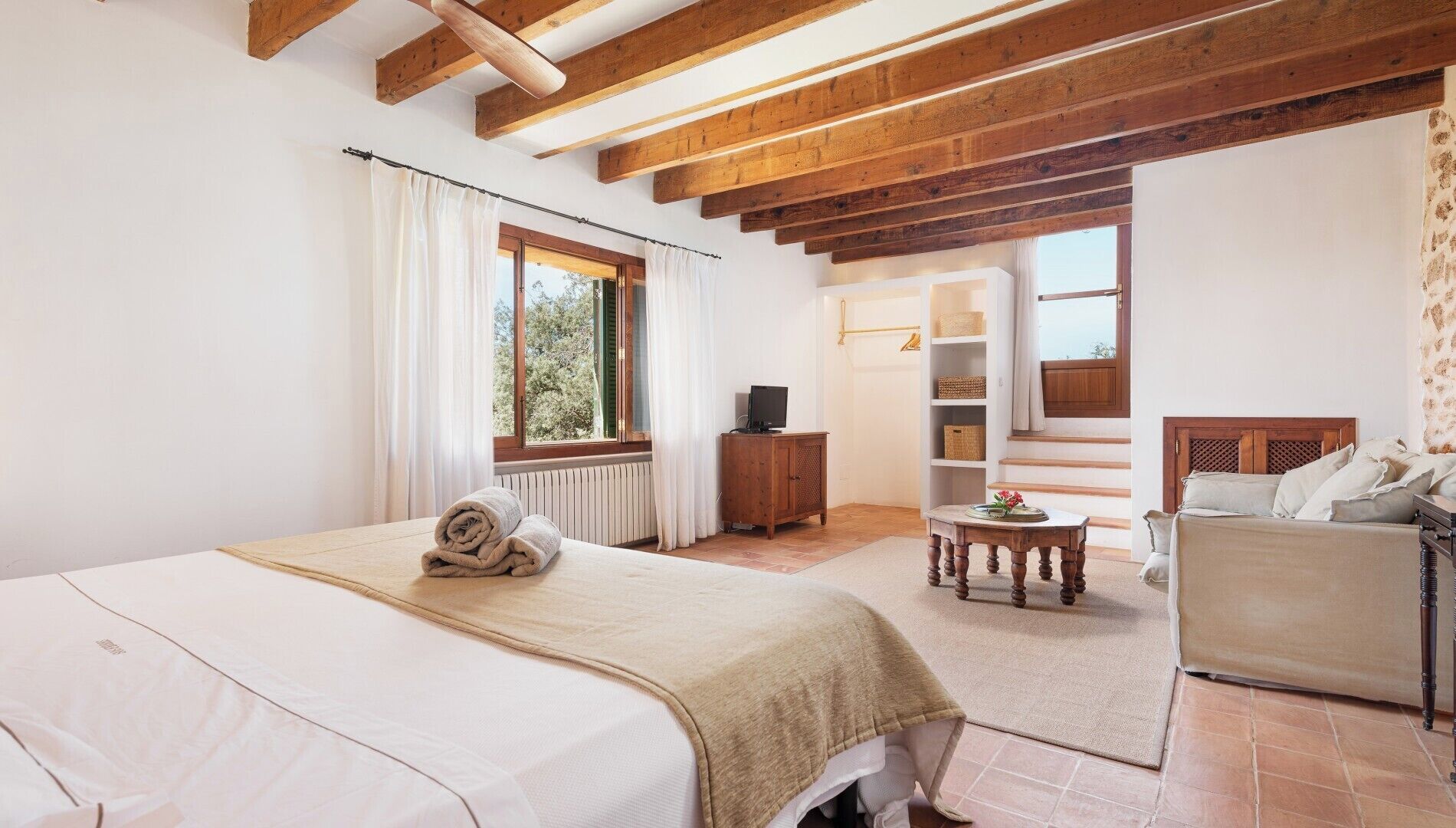 A bedroom with a bed, a wooden headboard, a small table, a sofa, and a window with curtains.