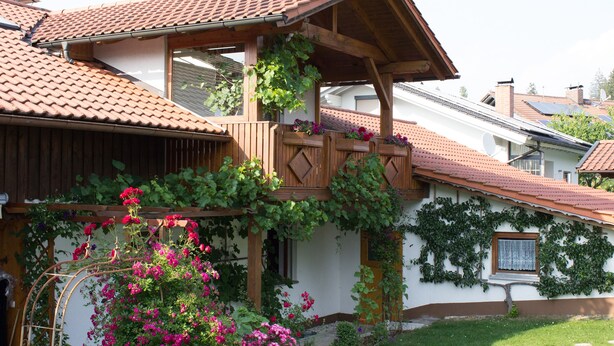 A house with a wooden balcony, a tiled roof, and a garden with flowering plants. - FeWo Konstandin (Bischofsmais)
