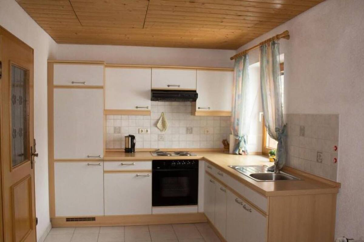 Cocina compacta con armarios blancos, horno negro, pileta de acero inoxidable y ventana con cortinas.