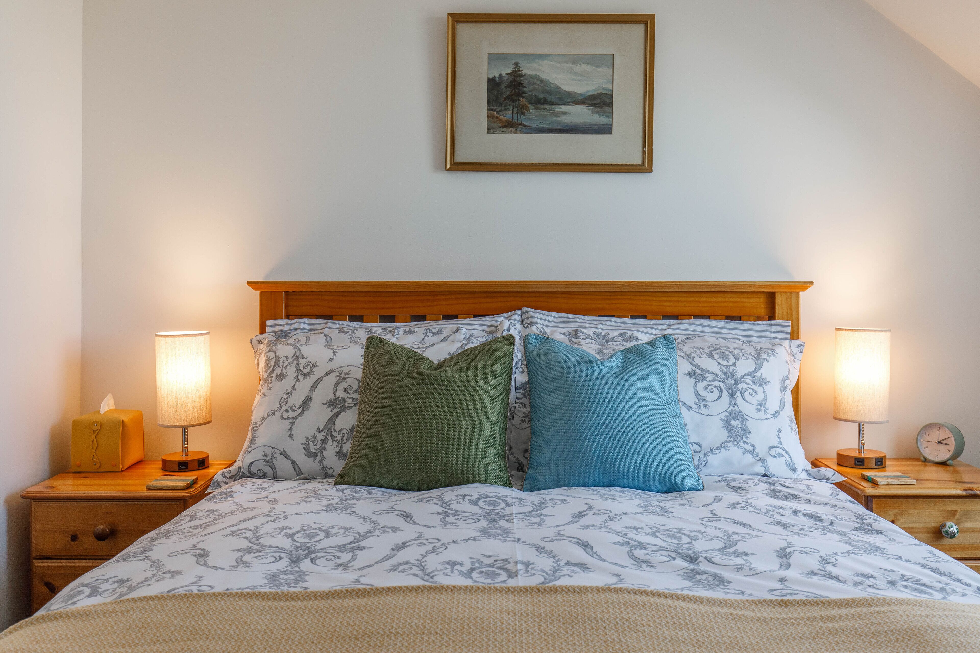 A neatly made bed with patterned bedding, two bedside lamps, a framed picture on the wall, and a wooden headboard.