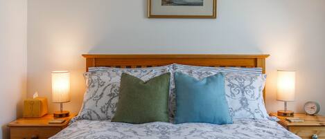 Una cama bien hecha con ropa de cama estampada, dos lámparas de noche, un cuadro en la pared y un cabecero de madera.