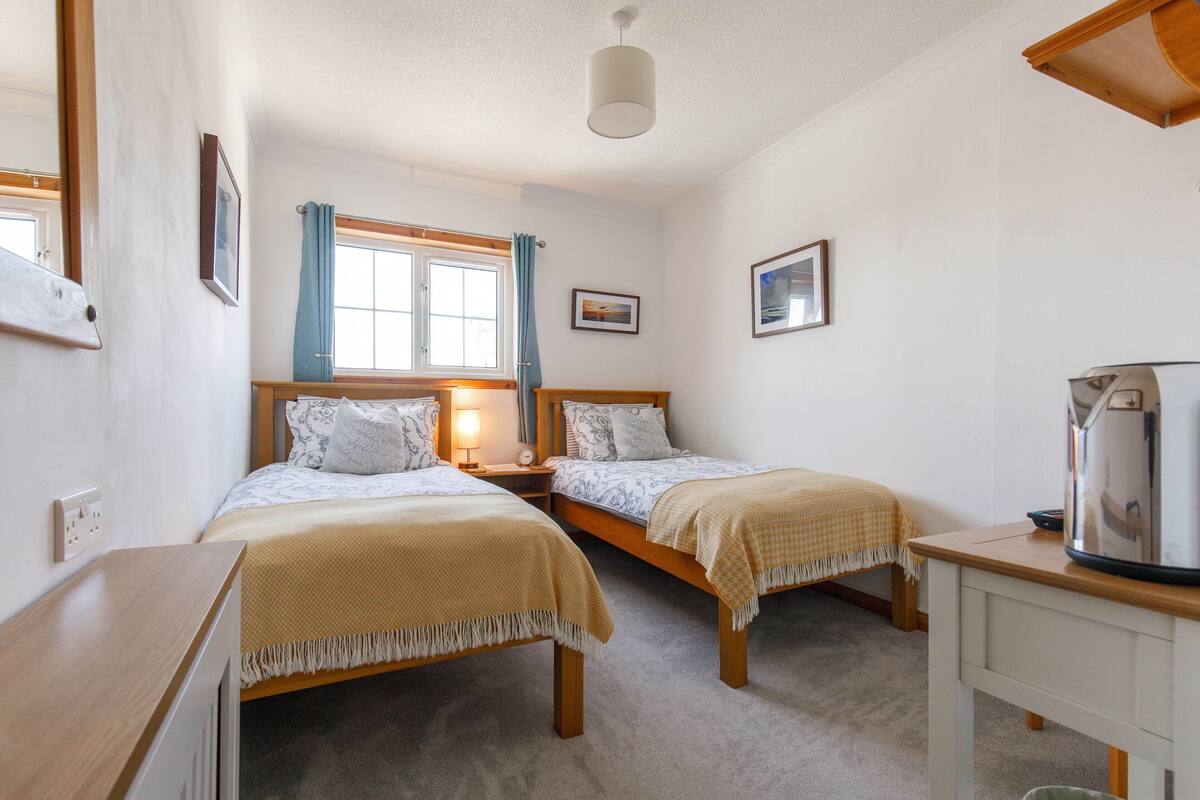 A bedroom with two beds, a window with curtains, a bedside table with a lamp, and a kettle on a table.