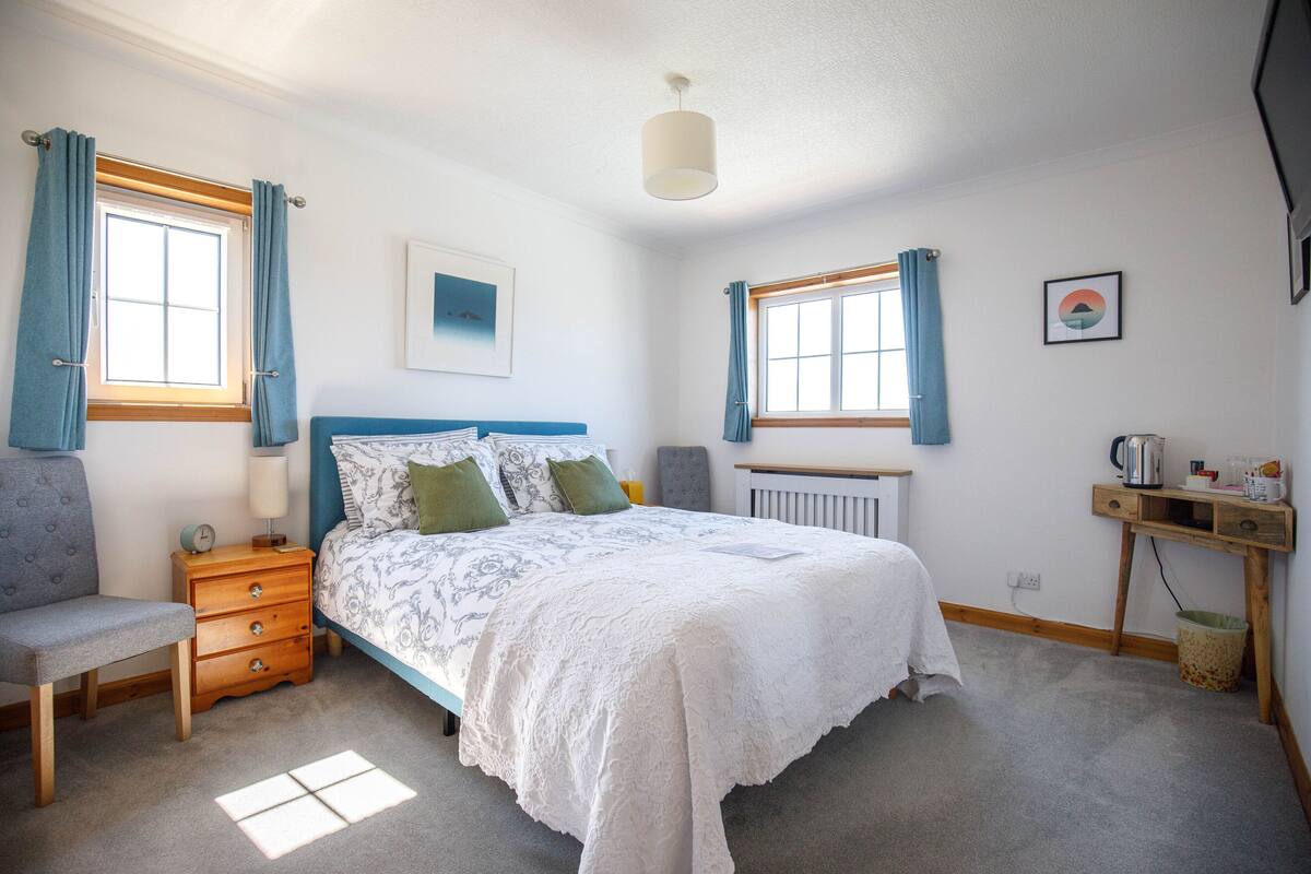 A bedroom with a bed, bedside table, lamp, chair, and a desk with a kettle.