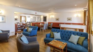 A spacious hotel lobby with a blue sofa, grey armchairs, a wooden coffee table, and a dining area with a red booth. - Cross Inn (Isle of Lewis)