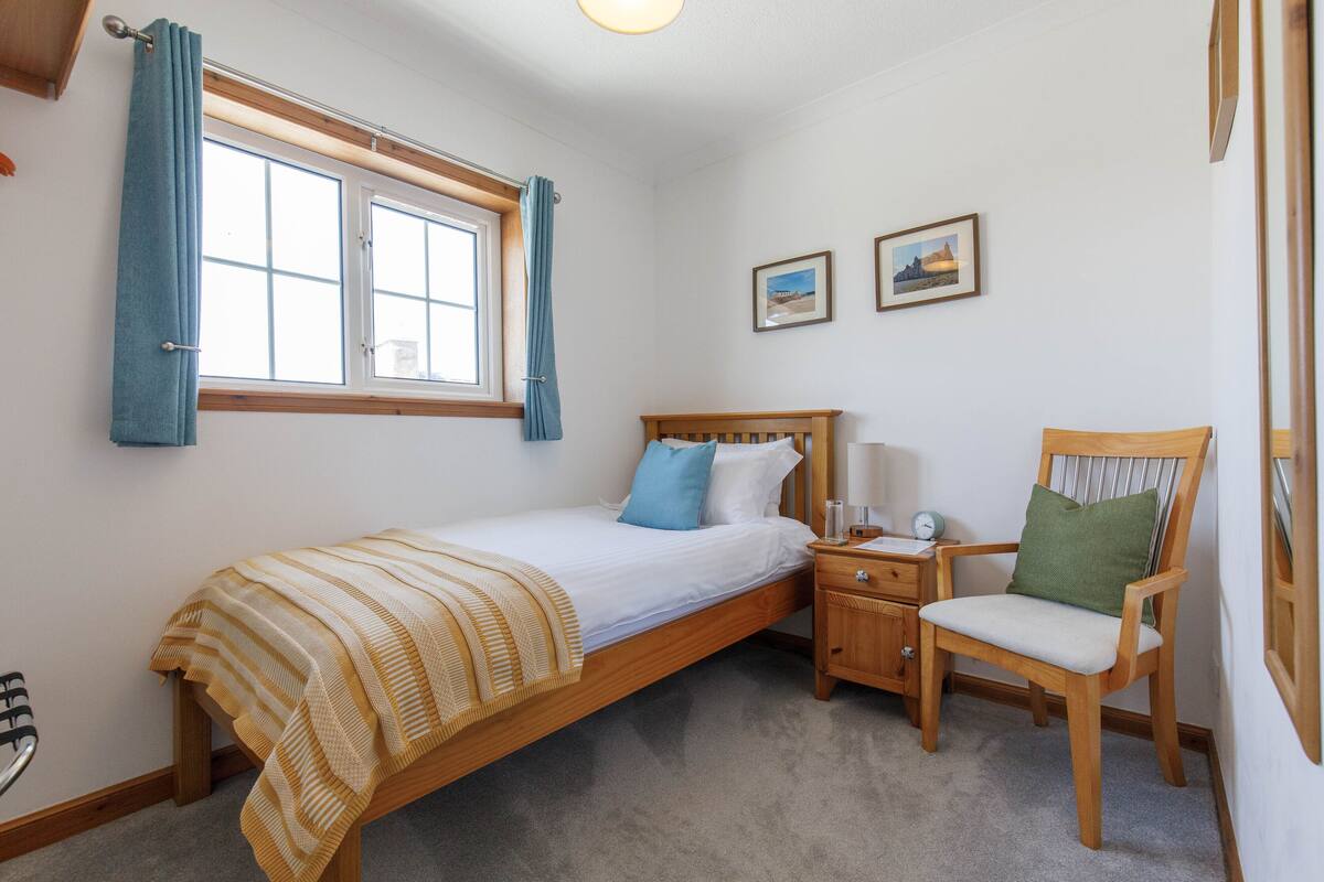 A bedroom with a single bed, a wooden nightstand, a chair, a window with curtains, and two framed pictures on the wall.