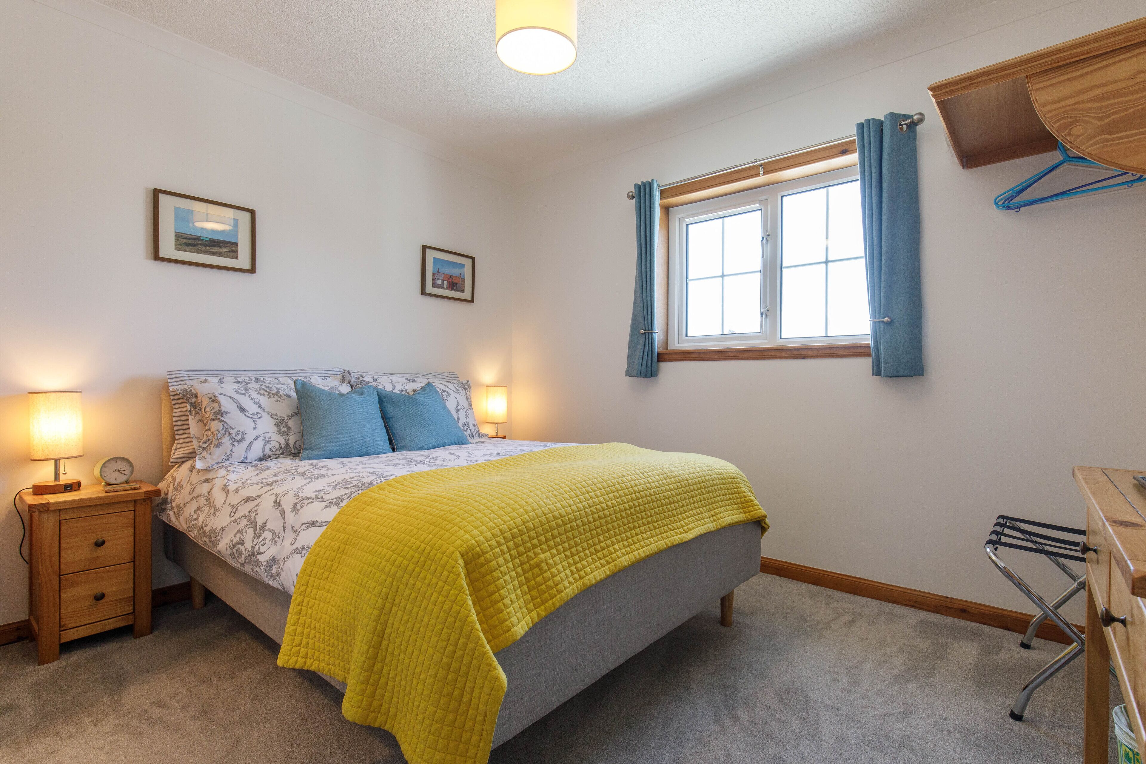 Un dormitorio con cama, lámparas de noche, una ventana con cortinas, una mesita de noche de madera y un tendedero fijado a la pared.