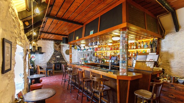 A rustic bar area with wooden bar stools, a counter with various bottles, and a stone wall. - Cross Inn (Isle of Lewis)