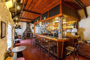 A rustic bar area with wooden bar stools, a counter with various bottles, and a stone wall. - Cross Inn (Isle of Lewis)