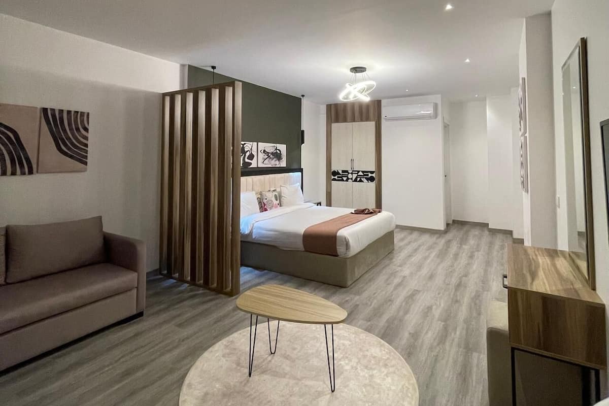 Moderni hotellihuone, jossa on sohva, pyöreä pöytä, puinen päätyosalla varustettu sänky ja suuri peili.