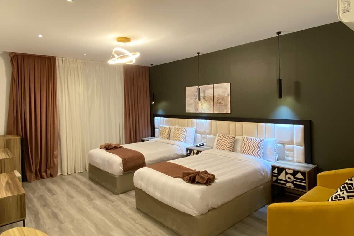 Moderni hotellihuone, jossa on kaksi sänkyä, keltainen nojatuoli, pieni pyöreä pöytä ja taulu seinällä.