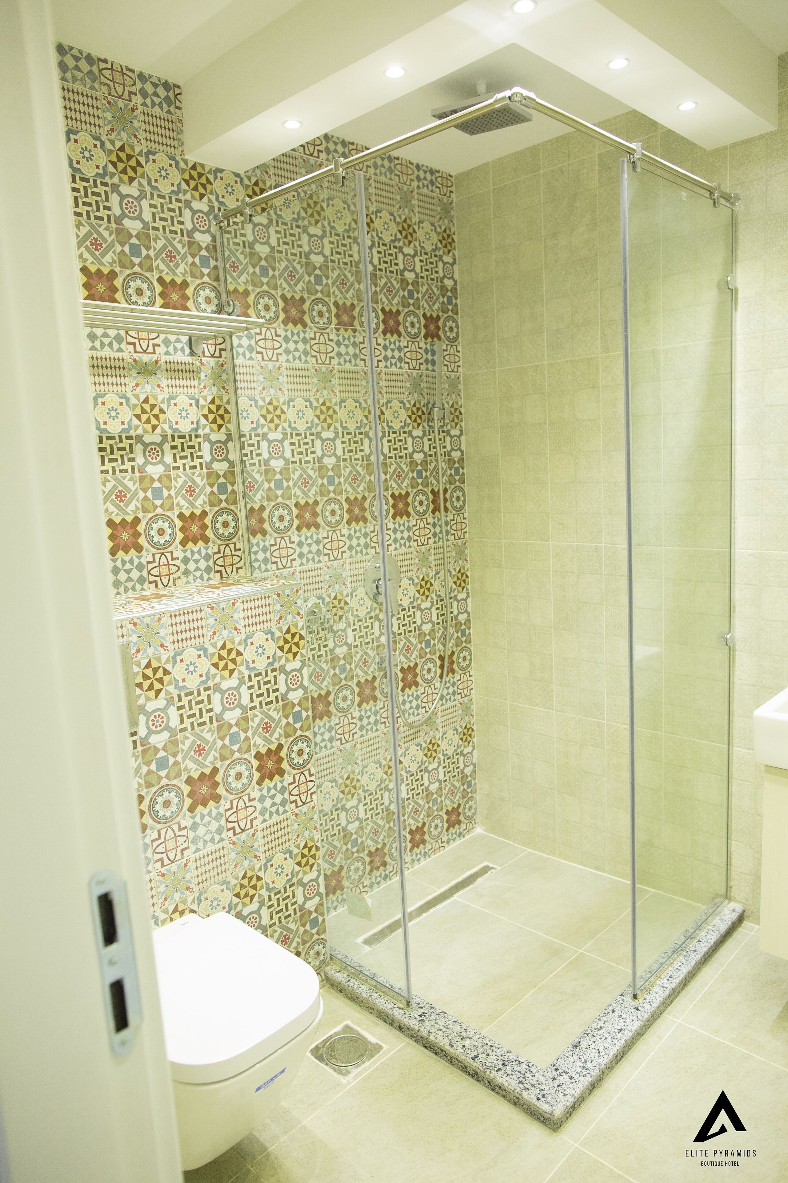 En bruser med glasdør, fliset væg med geometriske mønstre og et hvidt toilet.