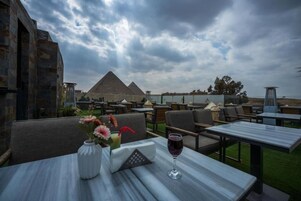 An outdoor dining area with tables and chairs, a vase with flowers, and a glass of red wine. In the background, there is a pyramid structure.