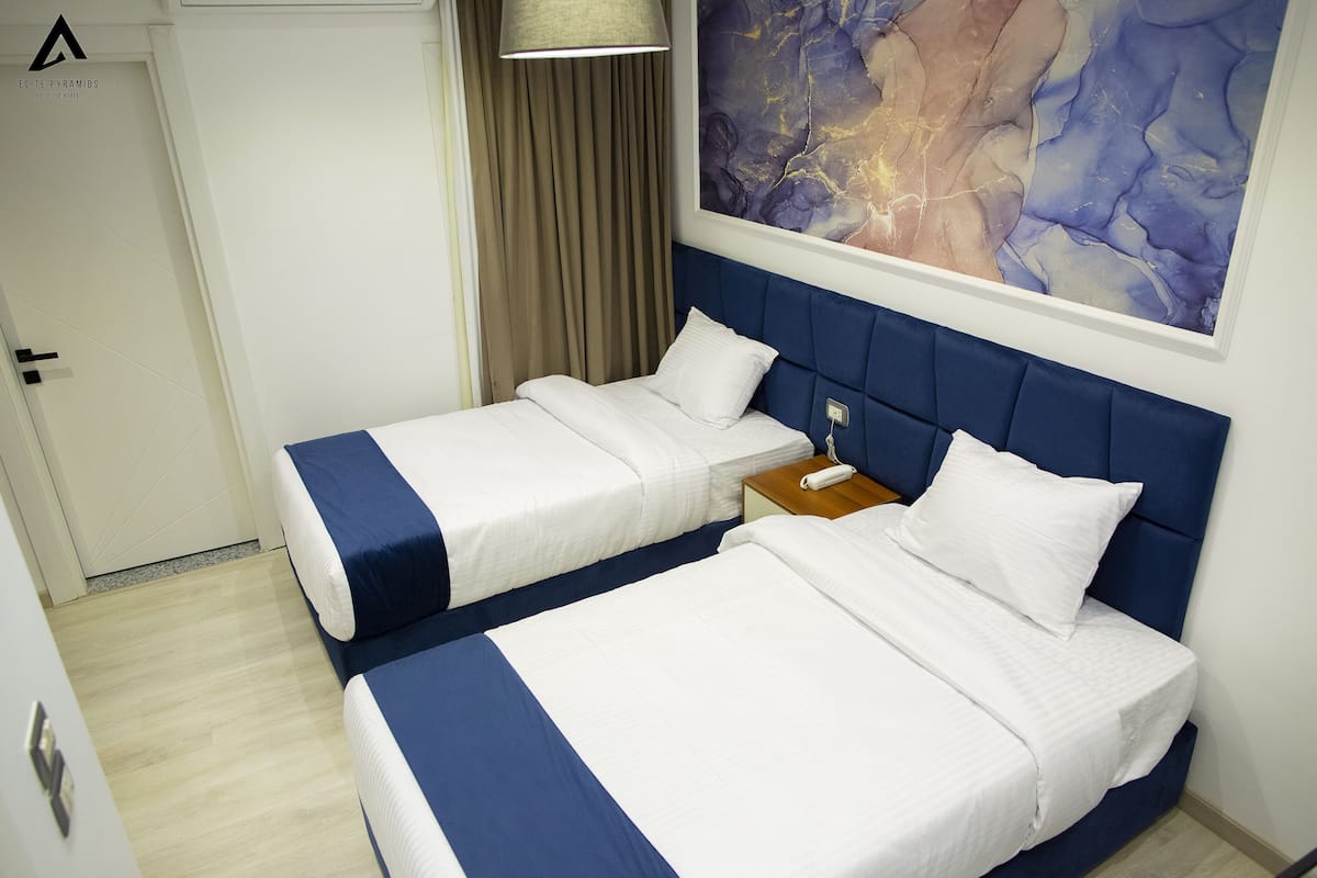 Hotellihuone, jossa on kaksi sänkyä, yöpöytä, lamppu ja taulu seinällä.