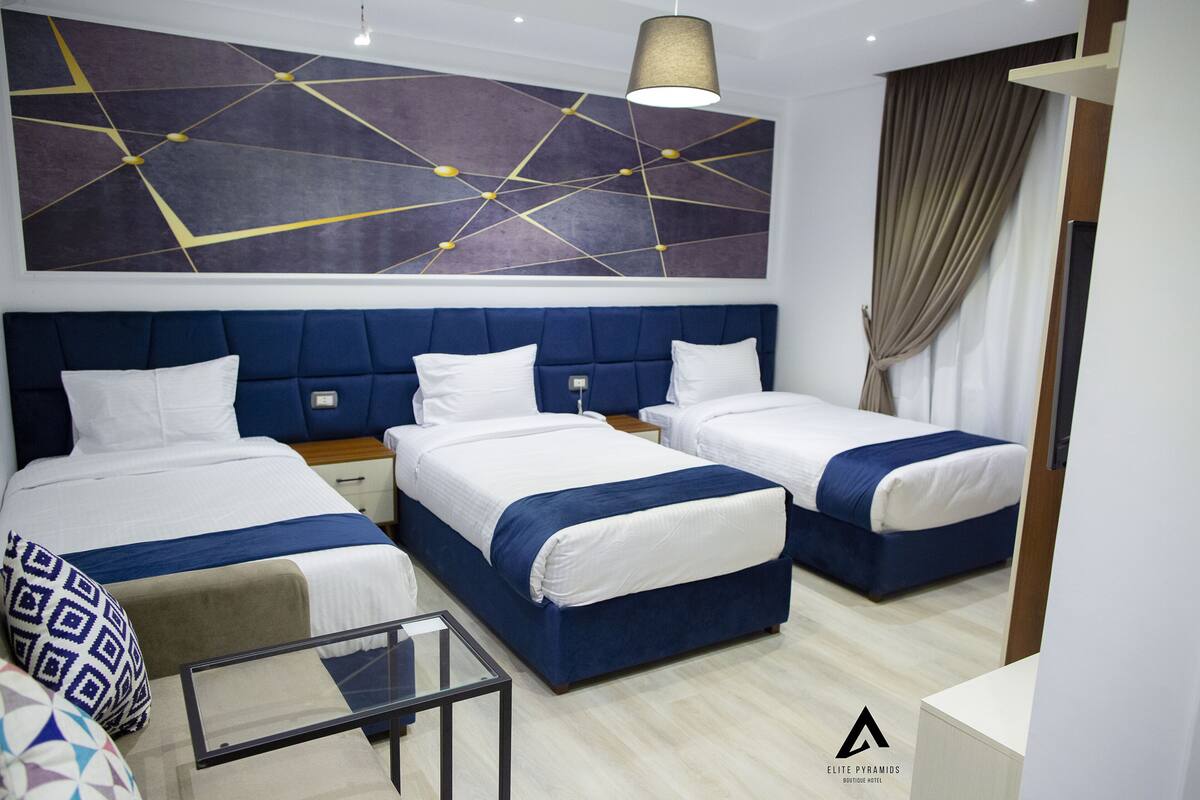 Hotellihuone, jossa on kolme sänkyä, seinällä moderni abstrakti maalaus ja lasinen sohvapöytä.