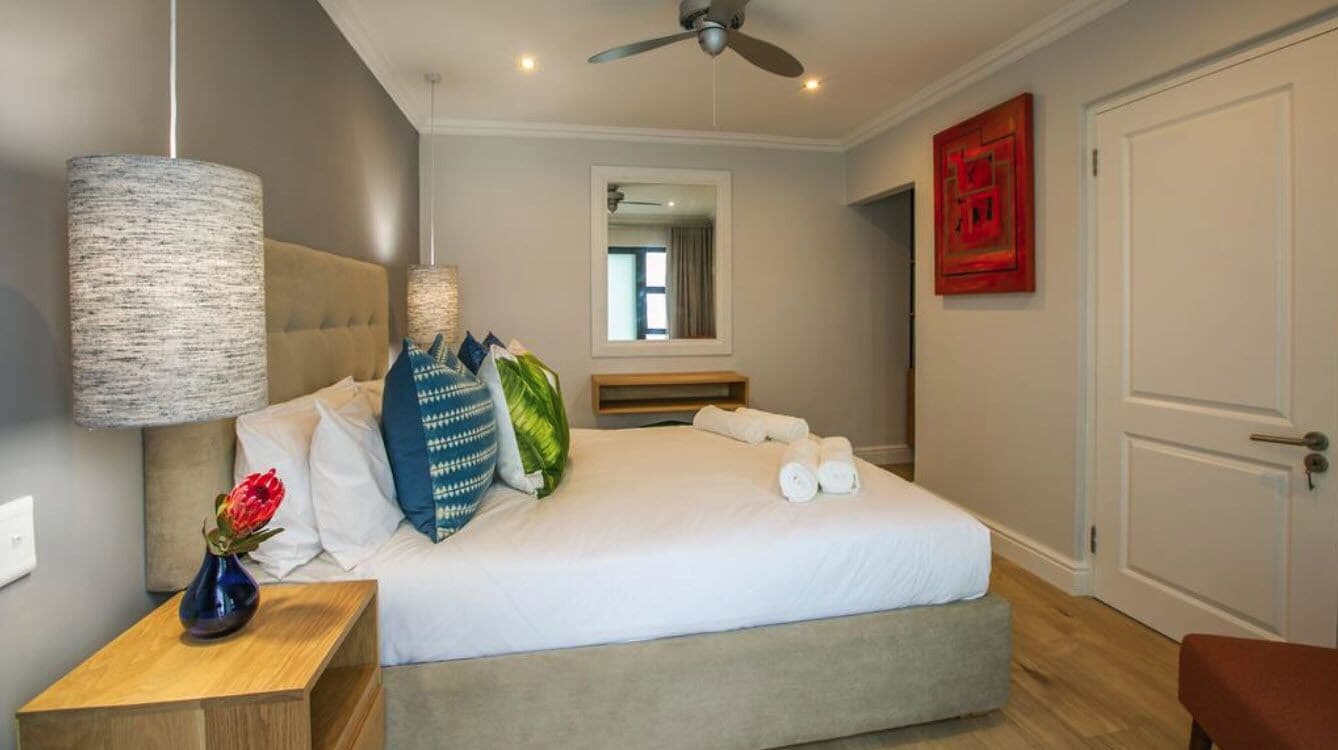 Uma cama bem arrumada com lençóis brancos, uma mesa de cabeceira de madeira com um vaso de flores, um ventilador de teto e uma porta com uma pintura abstrata vermelha.