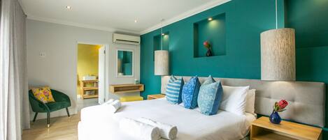 A modern hotel room with a large bed, a green armchair, a wooden nightstand, and a bathroom visible through an open door.