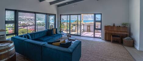 A spacious living room with a blue sectional sofa, large windows offering a view of the sea and a mountainous landscape, a wooden piano, and a rustic rug.