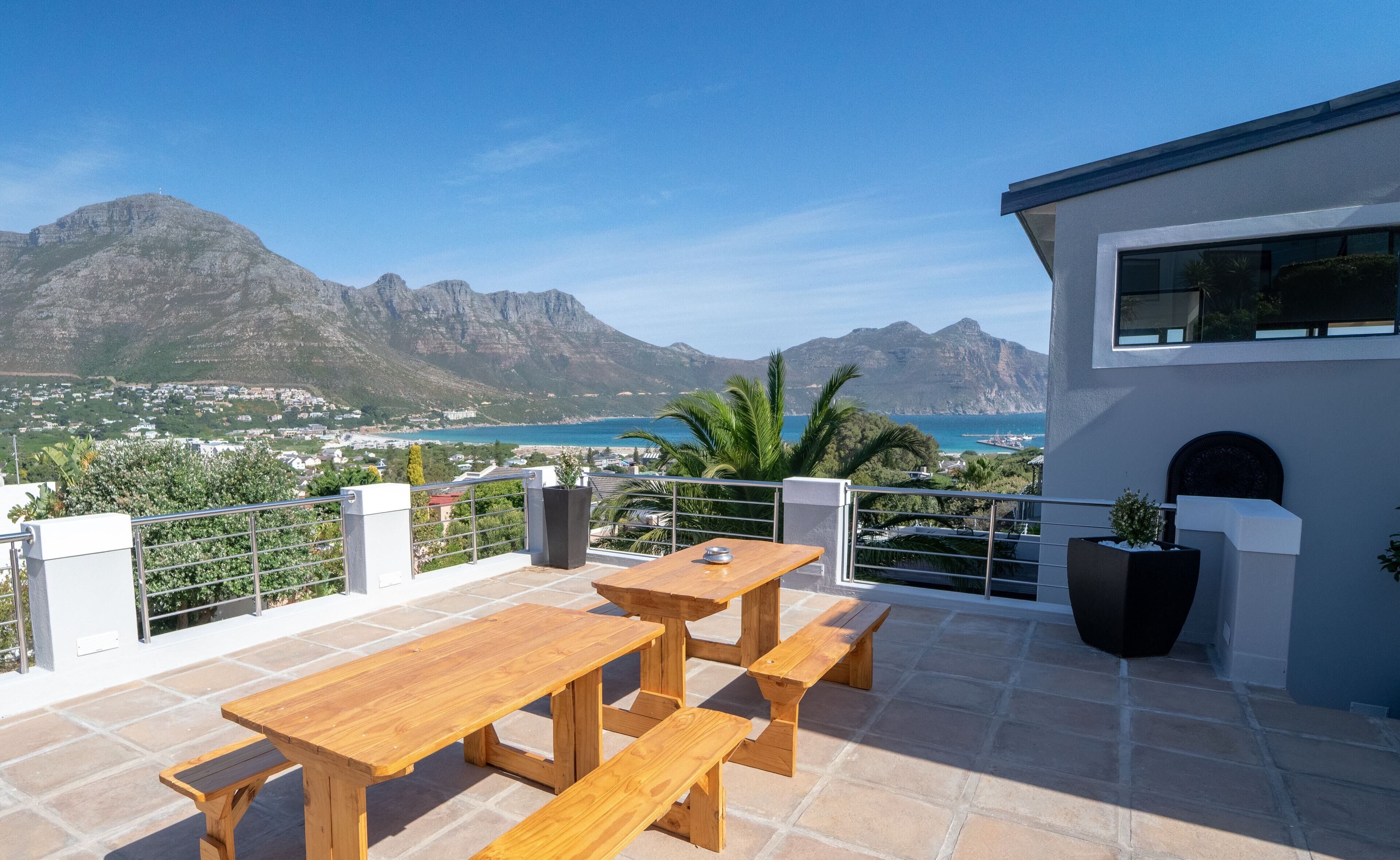 Una terraza con mesas y bancos de madera con vistas a un pueblo costero y montañas.