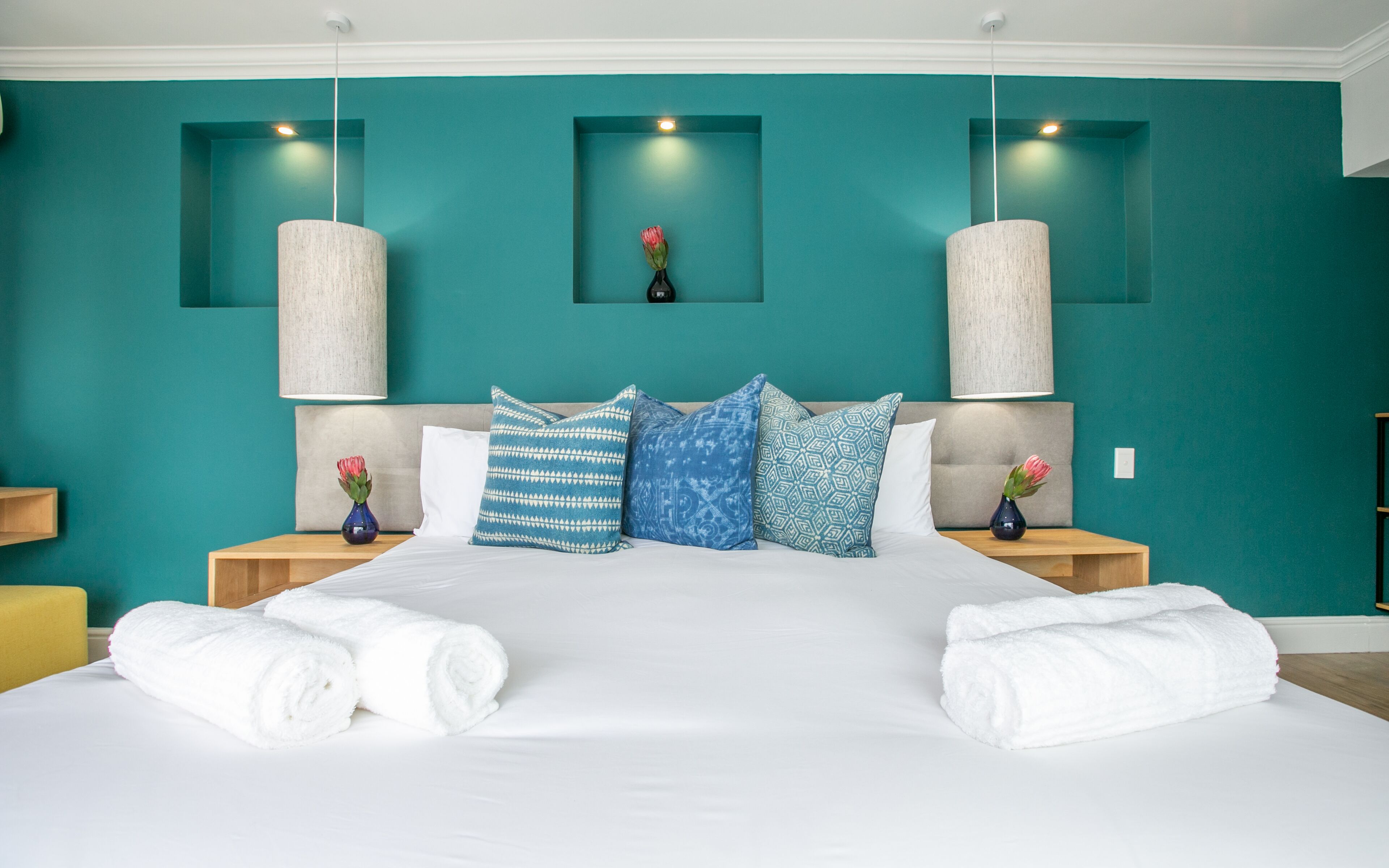 Una habitación de hotel moderna con paredes color turquesa, una cama grande con sábanas blancas, varios cojines estampados, dos mesitas de noche con lámparas y un pequeño florero con una flor en cada mesita.