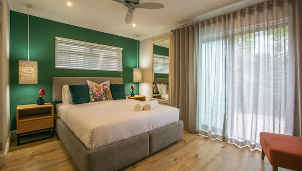 A bedroom with a large bed, two bedside tables, a ceiling fan, and a window with curtains. - Hout & About Guest House (Cape Town)