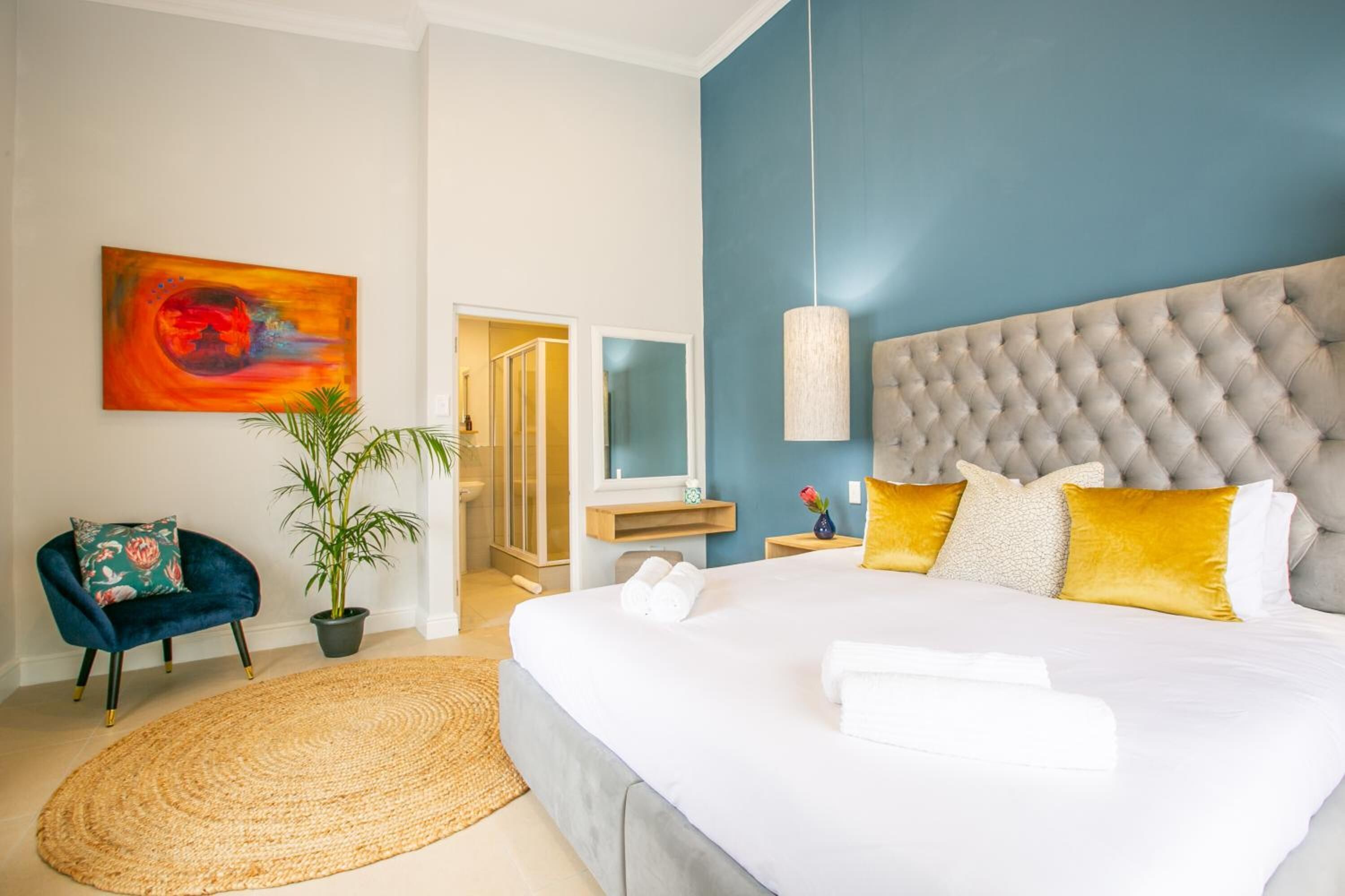 Une chambre moderne avec un grand lit, une tête de lit capitonnée, une plante en pot, un mur d’accent bleu et un tableau accroché au mur.