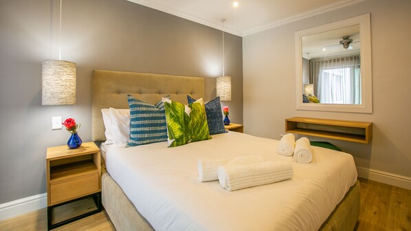 A neatly made bed with white linens and rolled towels, a headboard, and decorative pillows. There are bedside tables with lamps and a vase with flowers. A mirror hangs on the wall, and a wooden shelf is visible. - Hout & About Guest House (Cape Town)
