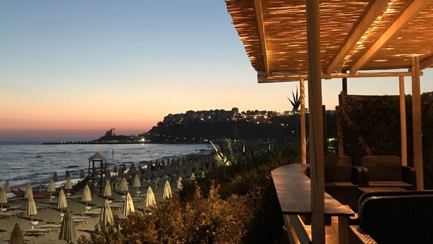 A beachside view with a covered seating area, umbrellas, and a view of the sea and distant cityscape. - Charming villa pool near sea to couple with stay in Italy (Itri)