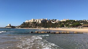 A coastal beach with clear waters, a sandy shore, and buildings on a hillside in the background. - Charming villa pool near sea to couple with stay in Italy (Itri)