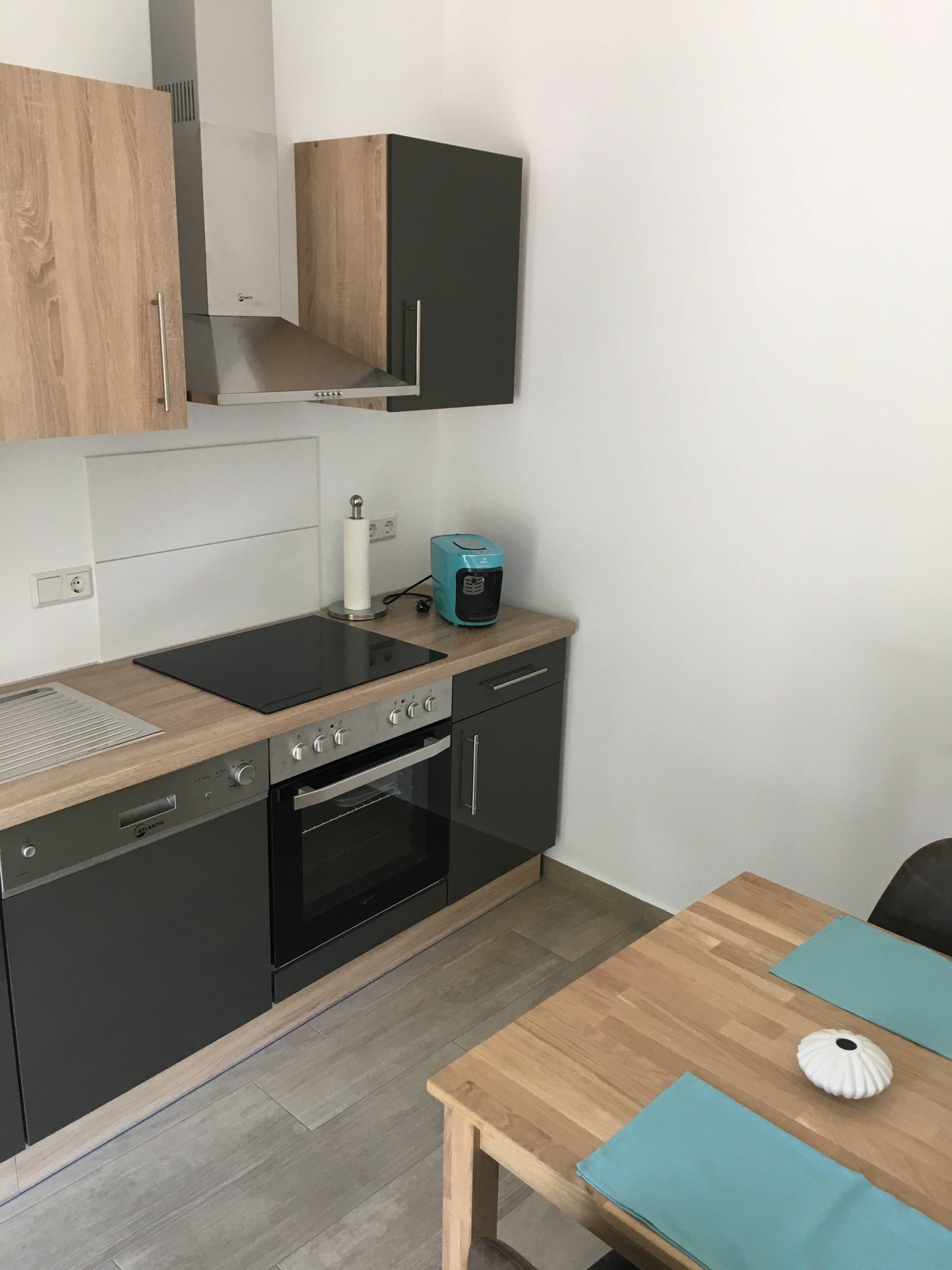 Une cuisine compacte avec un lave-vaisselle noir, un four encastré, une plaque de cuisson et une petite table agrémentée d’un set de table couleur turquoise.