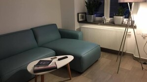 A living room with a teal sofa, a small round coffee table with books and a remote, a floor lamp, and a window with plants on the sill. - Apartments am Moselufer (Bruttig-Fankel)