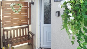 A wooden bench, a small table with drinks, a door with a nameplate, and a wall-mounted planter with flowers.