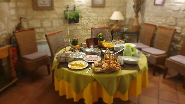 Una mesa de comedor preparada con diferentes platos y bebidas, rodeada de sillas, en una habitación con paredes de piedra y suelo de baldosas.