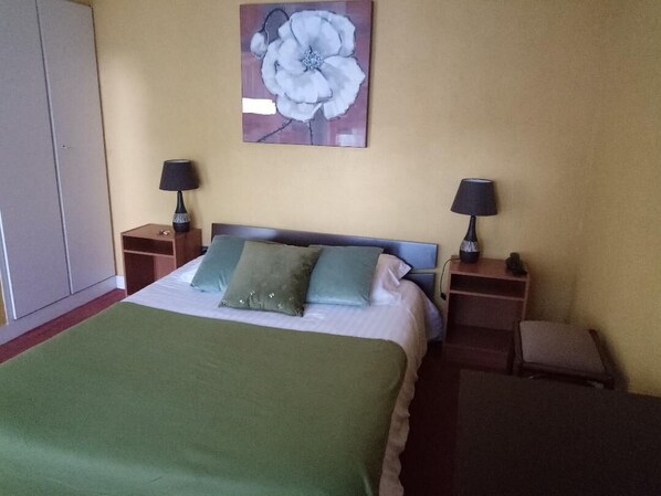 A neatly made bed with a green blanket, two bedside tables with lamps, and a wall-mounted flower painting. - L'Hostellerie De La Rivière (Creil)