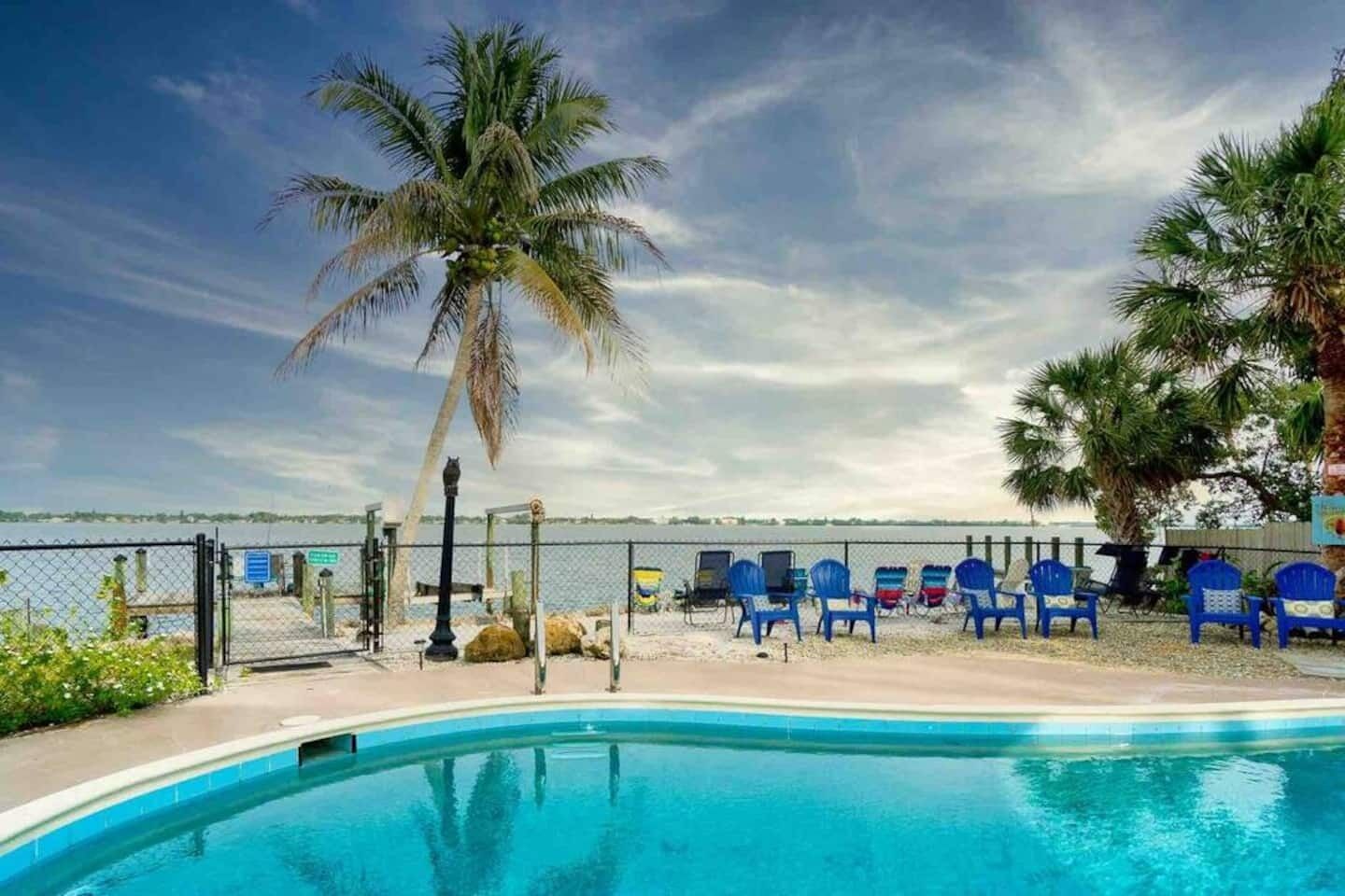 A pool area with blue chairs, a palm tree, and a fence overlooking a body of water.