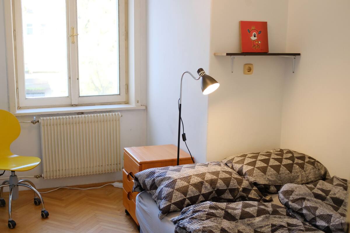 A small, well-lit room with a window, a desk with a chair, a bed with patterned bedding, and a shelf with a picture.