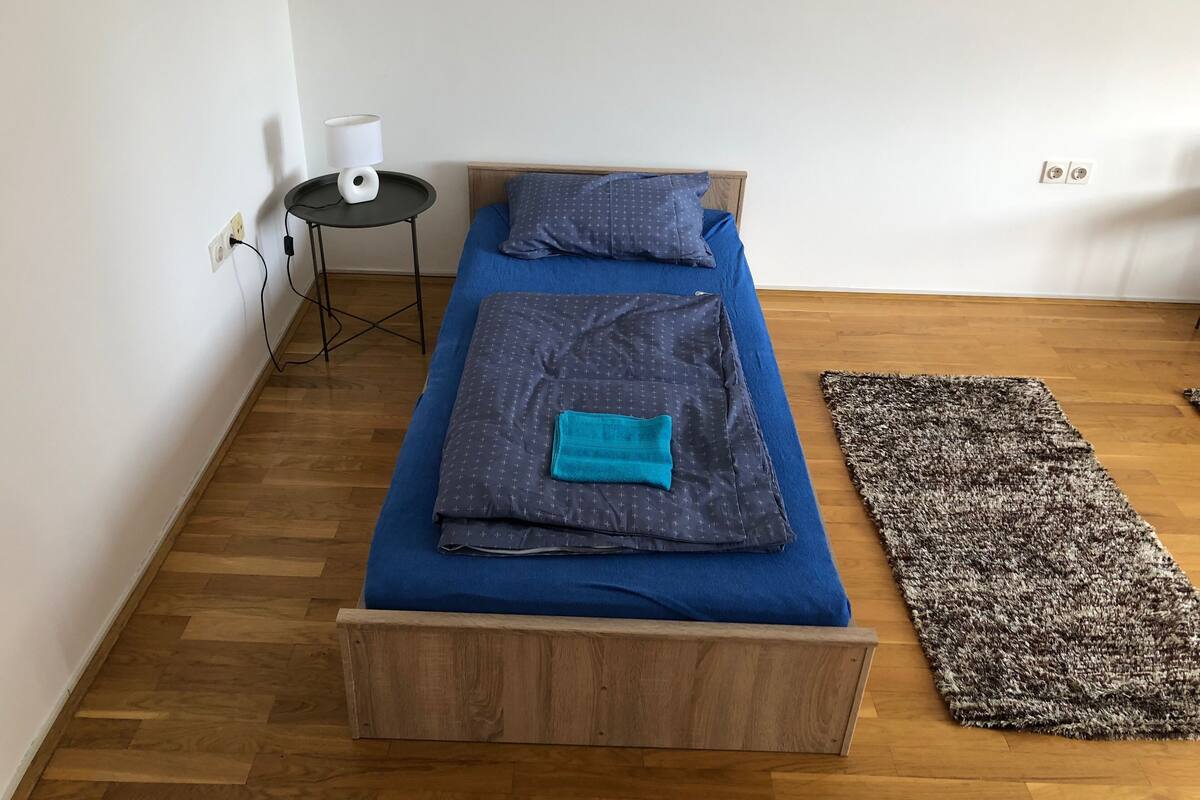 A single bed with a blue mattress and a matching blue headboard, a small table with a lamp, and a dark patterned rug on a wooden floor.