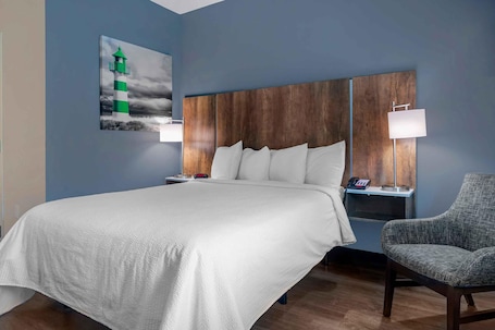 A hotel room with a large bed, a nightstand, a chair, a wall-mounted lamp, and a painting of a lighthouse.