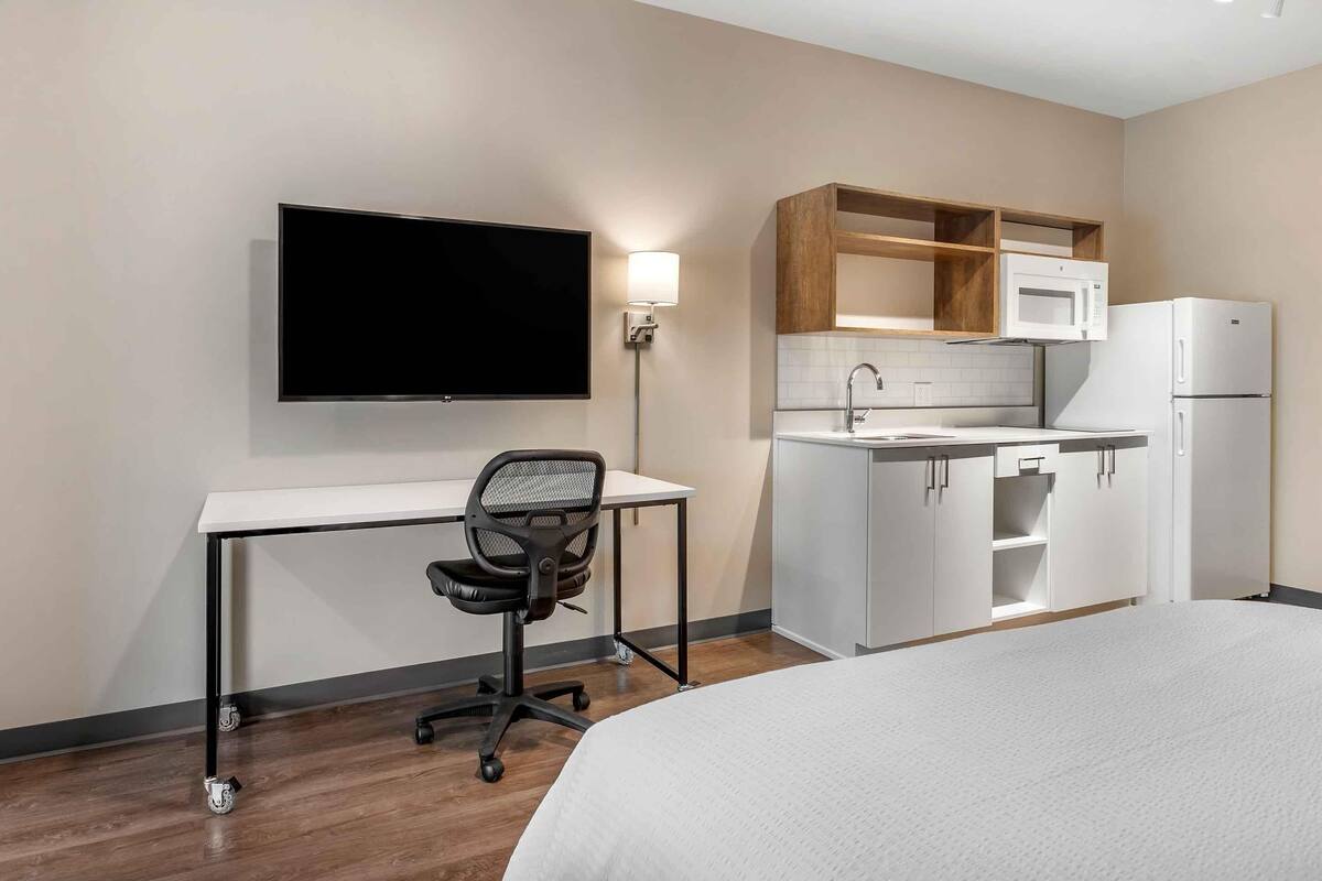 A hotel room with a bed, a desk with a chair, a flat-screen TV, a kitchenette with a microwave and refrigerator, and a wall-mounted light.