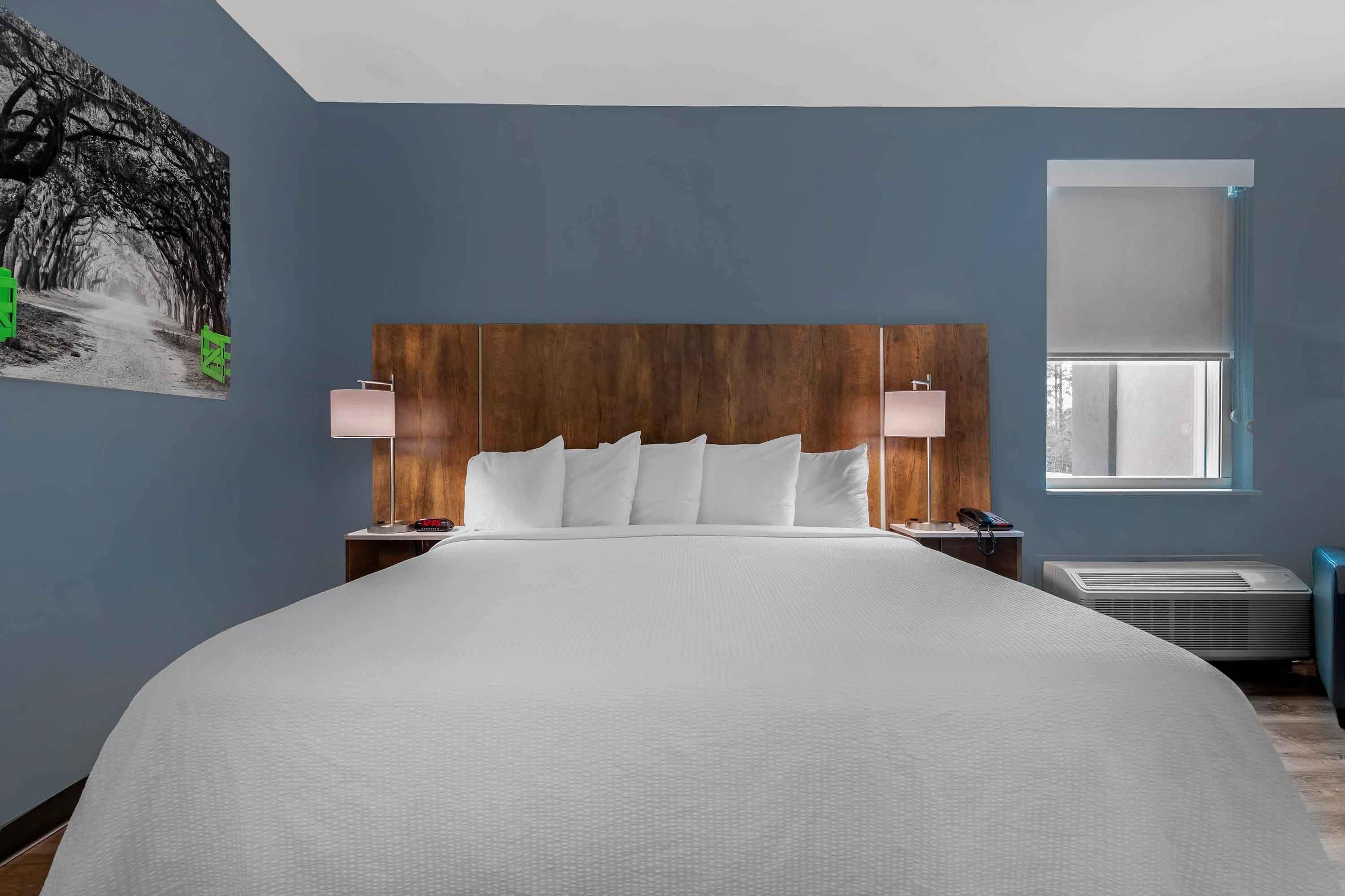 Habitación de hotel con una cama grande, cabecera de madera, dos mesitas de noche con lámparas, una ventana con persianas y un aire acondicionado instalado en la pared.