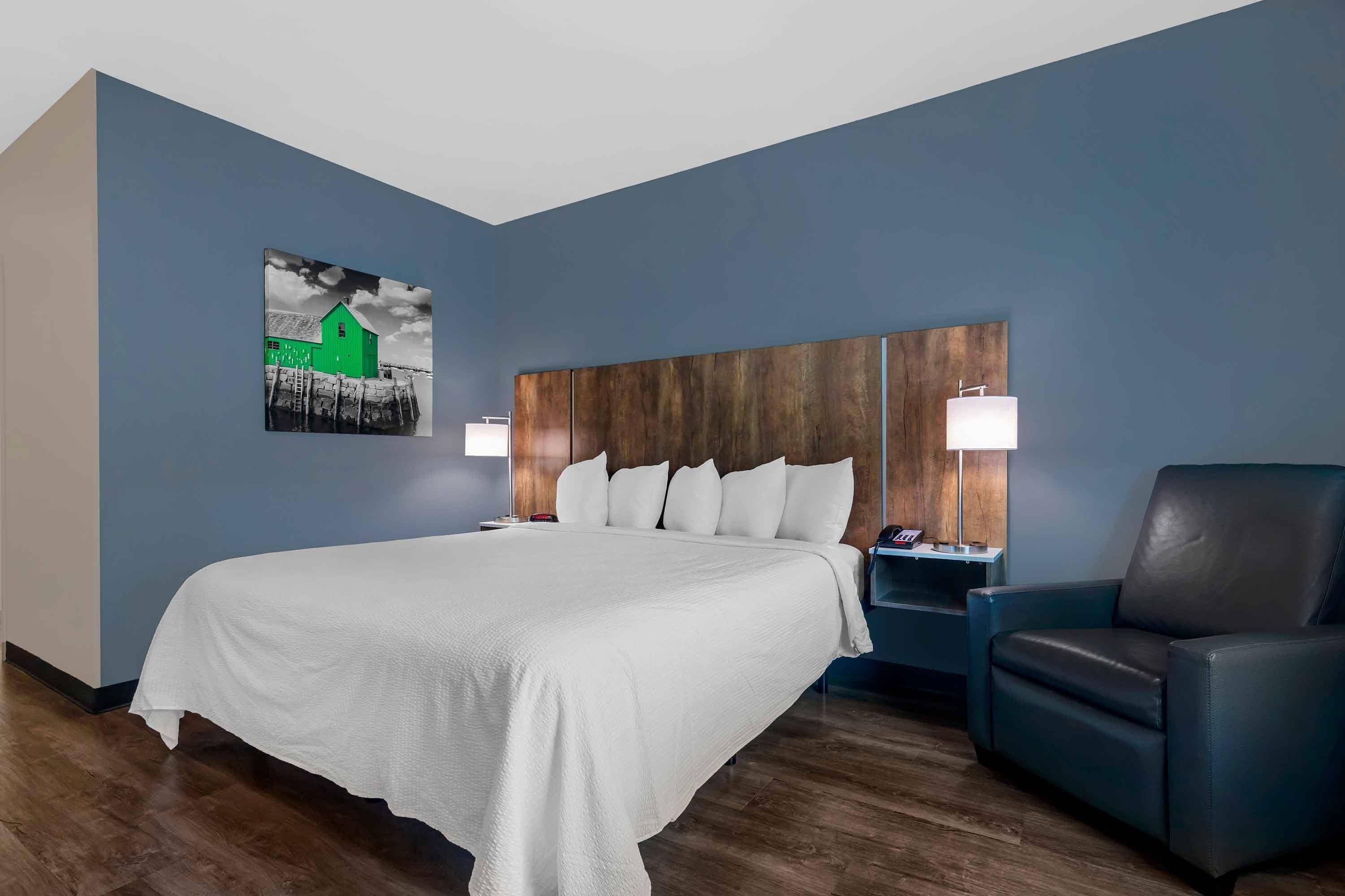 A hotel room with a large bed, a wooden headboard, two bedside tables with lamps, a black leather chair, and a wall-mounted artwork.