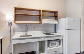 Una cocina compacta con encimera blanca, microondas integrado, refrigerador blanco y estantes de madera sobre la encimera.