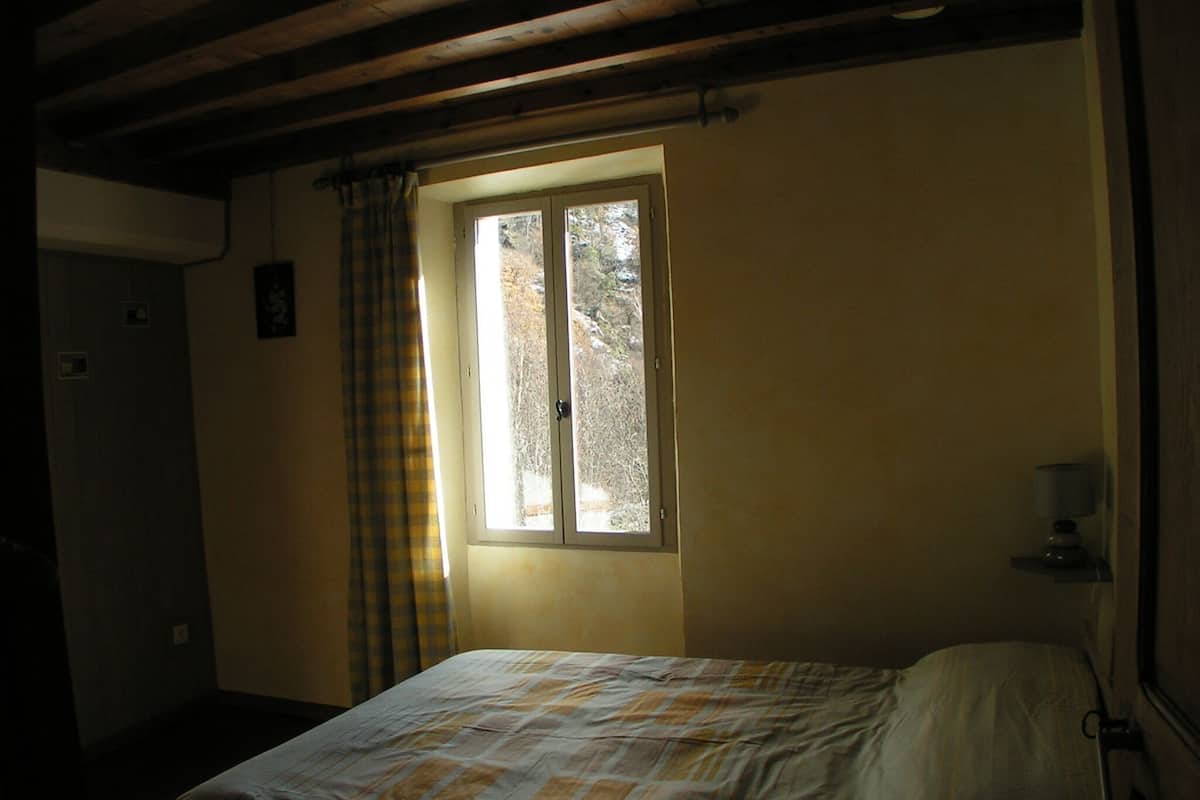Ein Zimmer mit einem Bett, einem Fenster mit Vorhängen, einem Nachttisch mit einer Lampe und einer Tür.