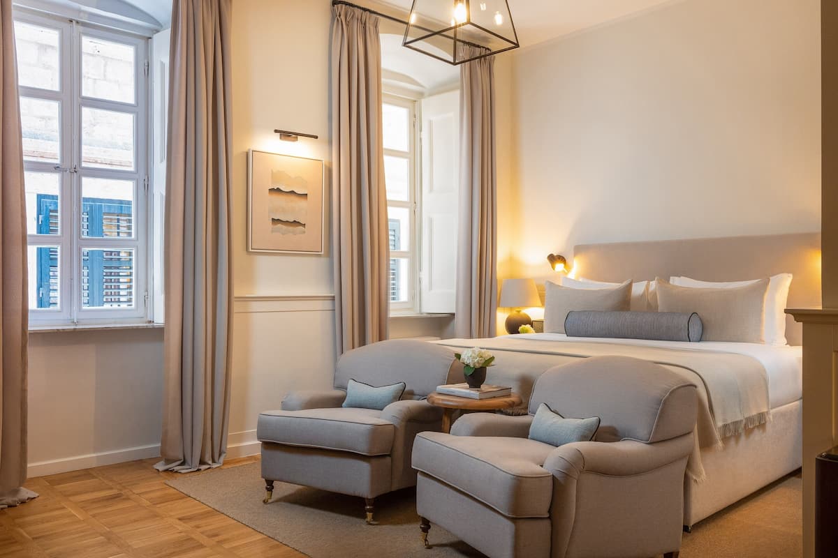 A bedroom with a large bed, two armchairs, a small table with a book and flowers, and a window with curtains.