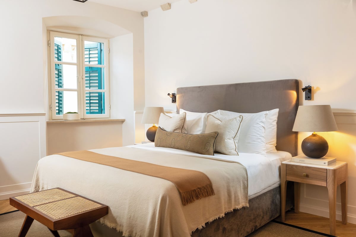 A neatly made bed with a headboard, two bedside tables with lamps, a window with shutters, and a small wooden stool.
