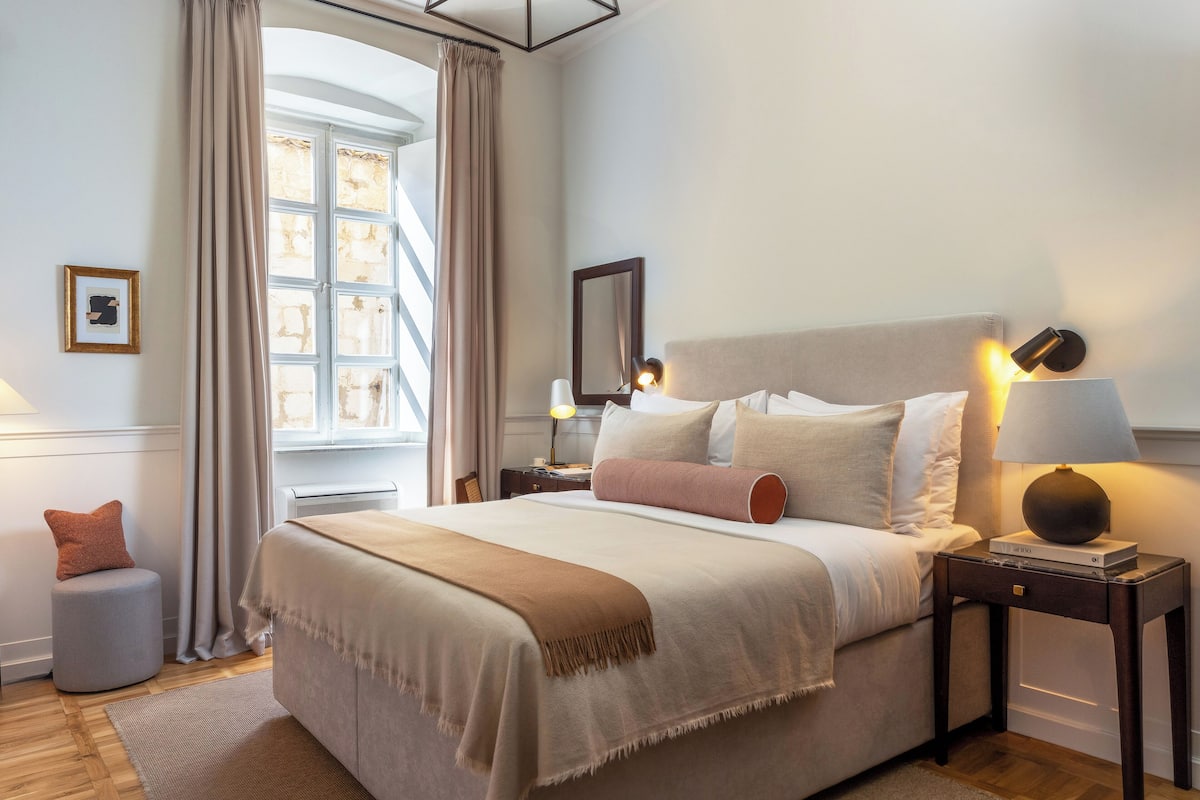 A neatly arranged bedroom with a large bed, a bedside table with a lamp, a small stool, and a window with curtains.
