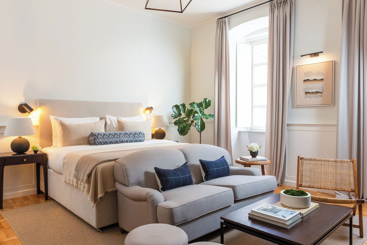 A modern hotel room with a large bed, a sofa, a coffee table, and a chair. There is a window with curtains, a plant, and a wall-mounted lamp.
