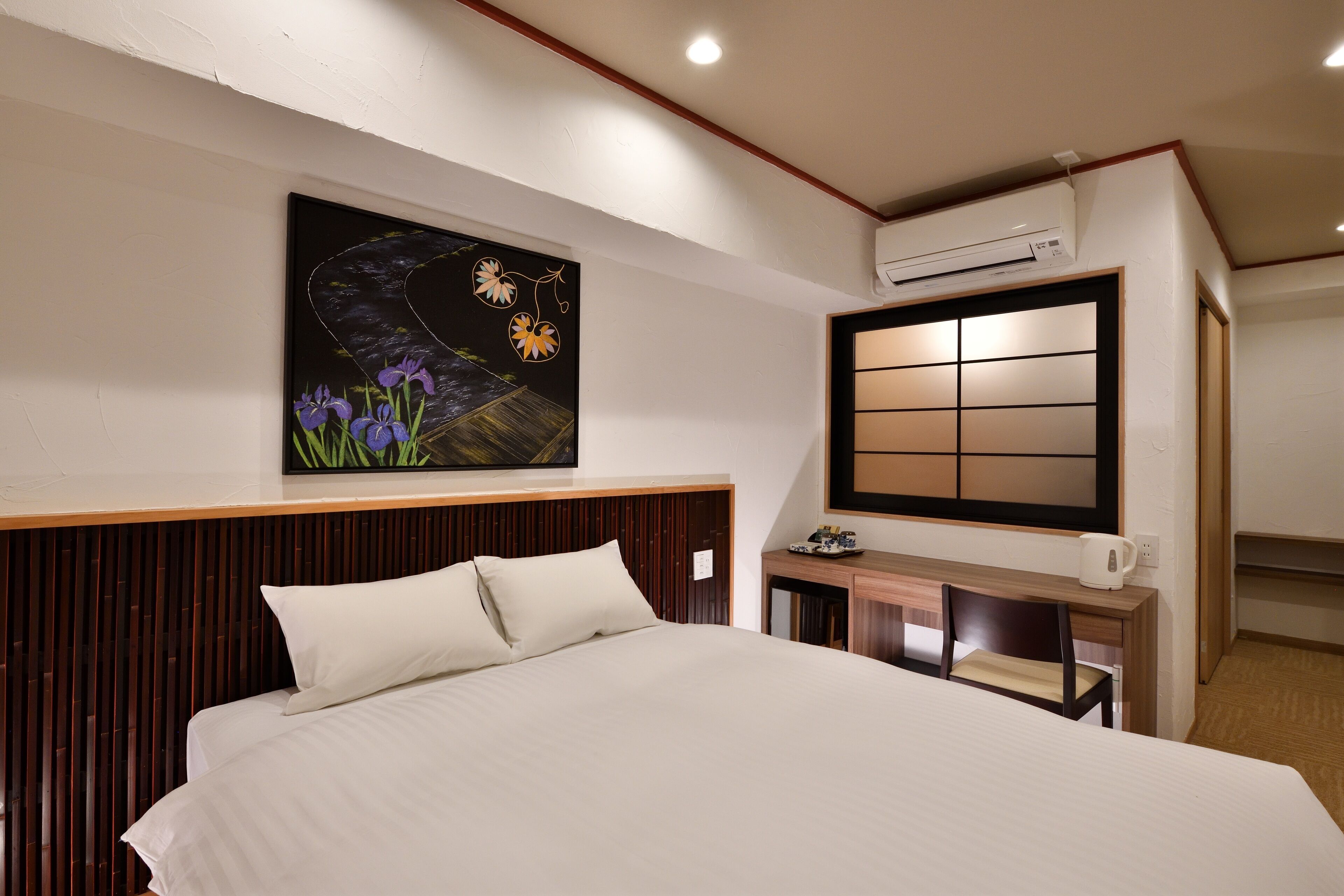 Habitación de hotel con una cama grande, un escritorio con silla, aire acondicionado de pared y un cuadro de flores.